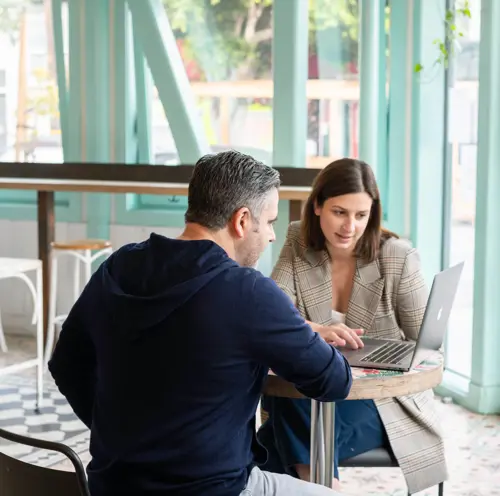 A man and woman sat at a round table in a cafe looking at a laptop.